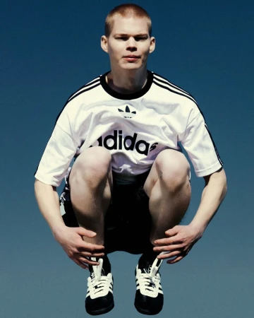 Athletic man in black-and-white Adidas shirt and shoes jumps against a vivid blue sky backdrop, showing sportswear and energy.