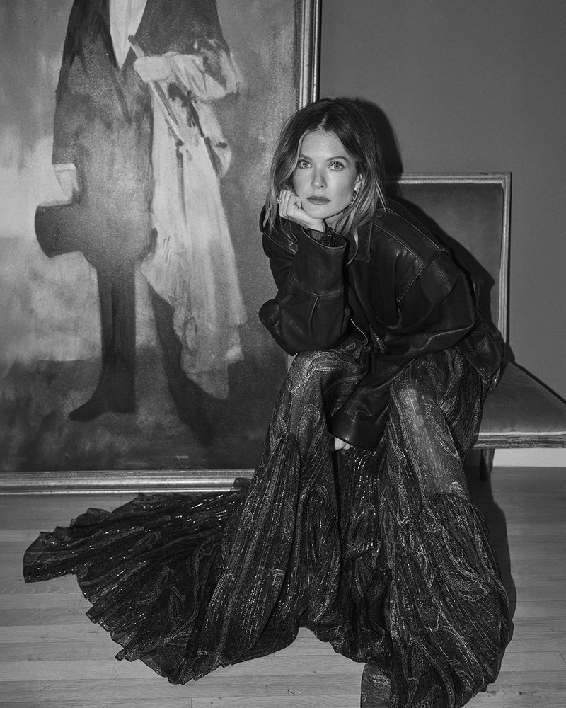 Actor Meghann Fahy wearing an oversized leather jacket and a voluminous shimmering gown, posed beside a large framed painting.
