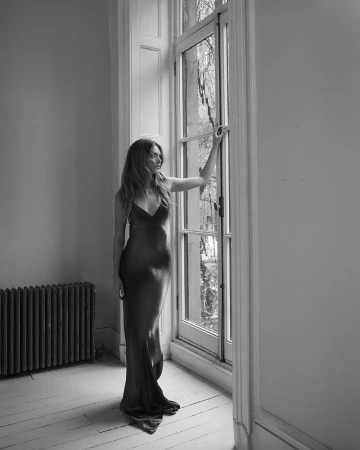 A woman in a long dress stands near a window in a room with white walls and wood floors, looking outside during daylight, in black and white.