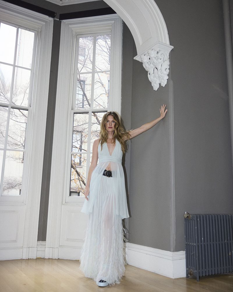 A full-length portrait of a woman with long hair, wearing a light blue dress and sneakers, standing near a window in a room with gray walls.
