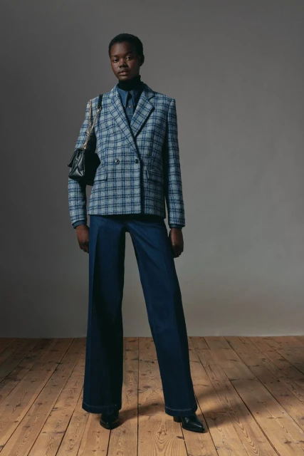 A full-length studio shot of a Black woman wearing a plaid blazer, a button-down shirt, and wide-leg jeans, with a black shoulder bag.