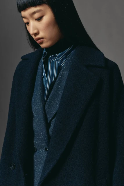 Asian woman wearing a dark blue wool coat over a pinstripe shirt and vest poses indoors against a dark grey backdrop.