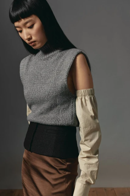 Adult woman in studio wearing a grey knit vest over a beige shirt and a brown skirt, looking downward in a waist-up shot.
