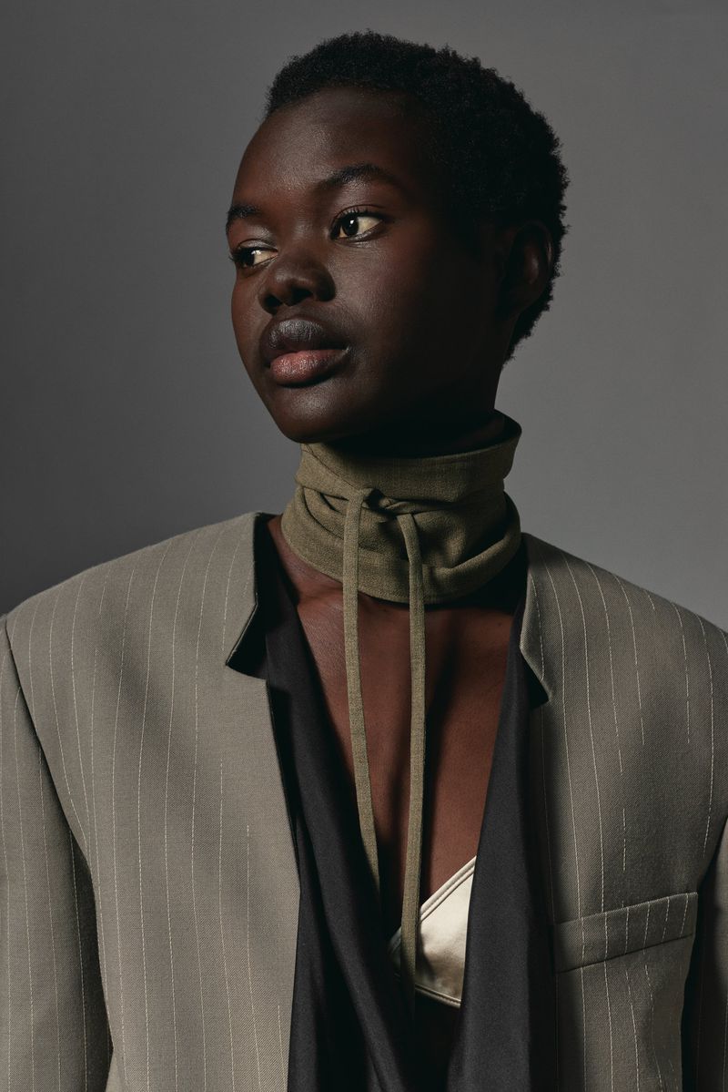 Striking portrait of an African American woman in a blazer, wearing a unique collar, against a neutral grey background. Fashion editorial.