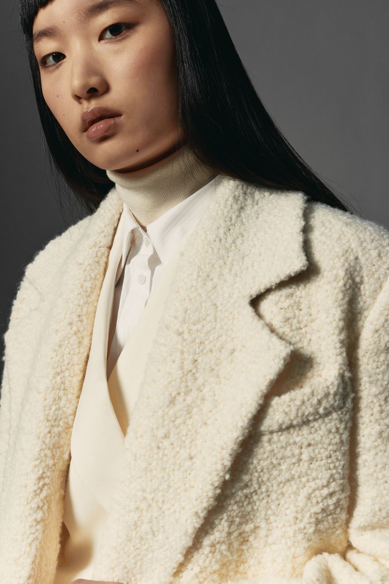 Portrait of a young Asian woman wearing a light-beige textured coat, a white shirt, and a light-beige turtleneck sweater.