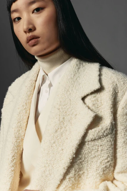 Portrait of a young Asian woman wearing a light-beige textured coat, a white shirt, and a light-beige turtleneck sweater.