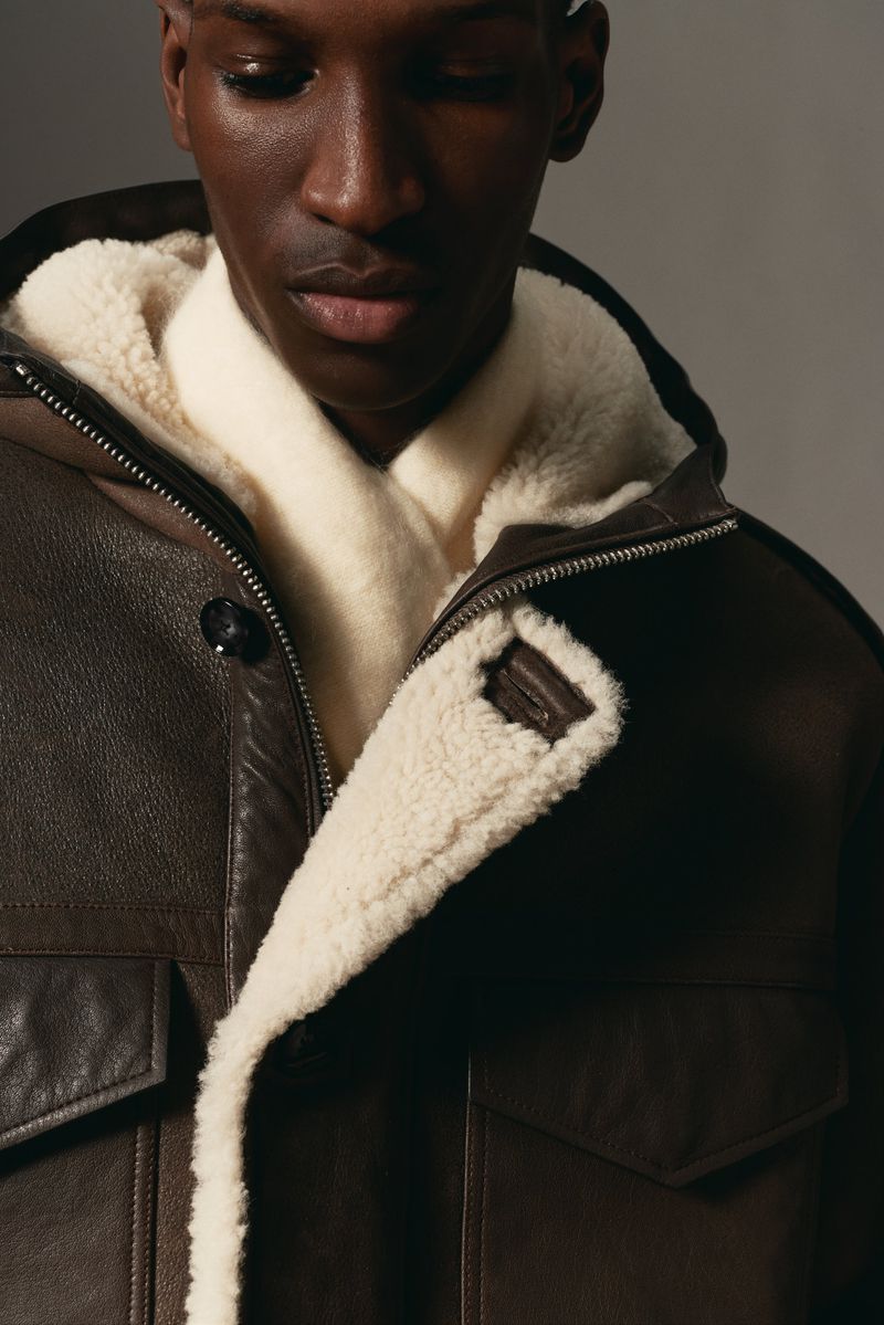 Close-up of an adult Black male wearing a brown leather jacket with a white shearling lining, looking downward in a studio setting.
