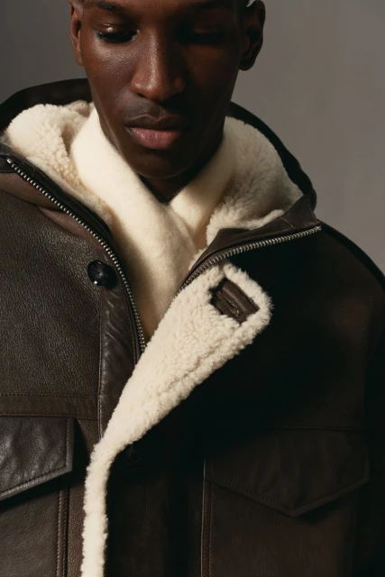 Close-up of an adult Black male wearing a brown leather jacket with a white shearling lining, looking downward in a studio setting.