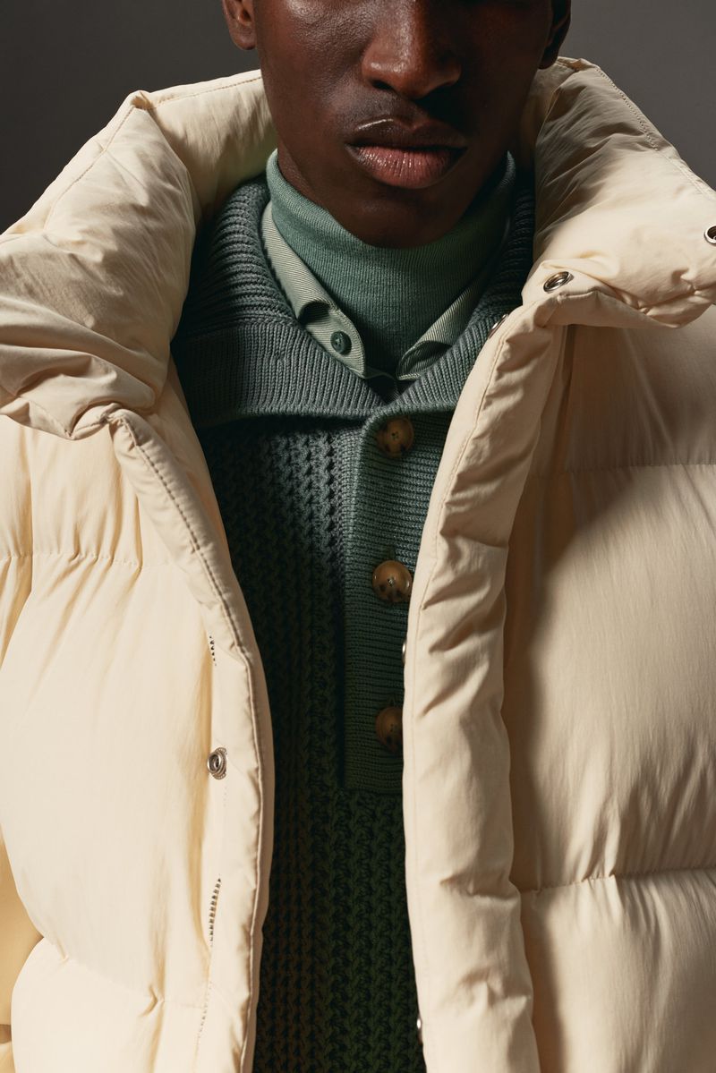 Close-up shot of a male model in a cream-colored puffer jacket with a ribbed, buttoned sweater, showcasing fashion and textures.