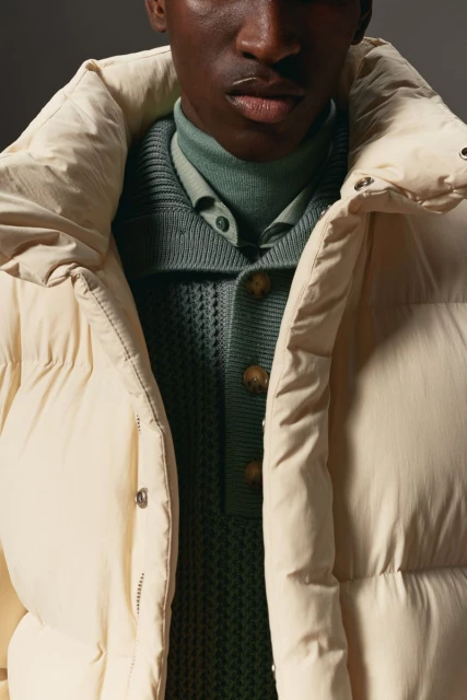 Close-up shot of a male model in a cream-colored puffer jacket with a ribbed, buttoned sweater, showcasing fashion and textures.