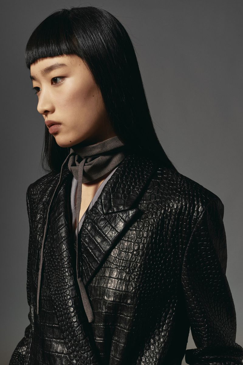 Portrait of a young Asian woman with black hair and a crocodile-skin blazer on a gray background in a fashion editorial shot.