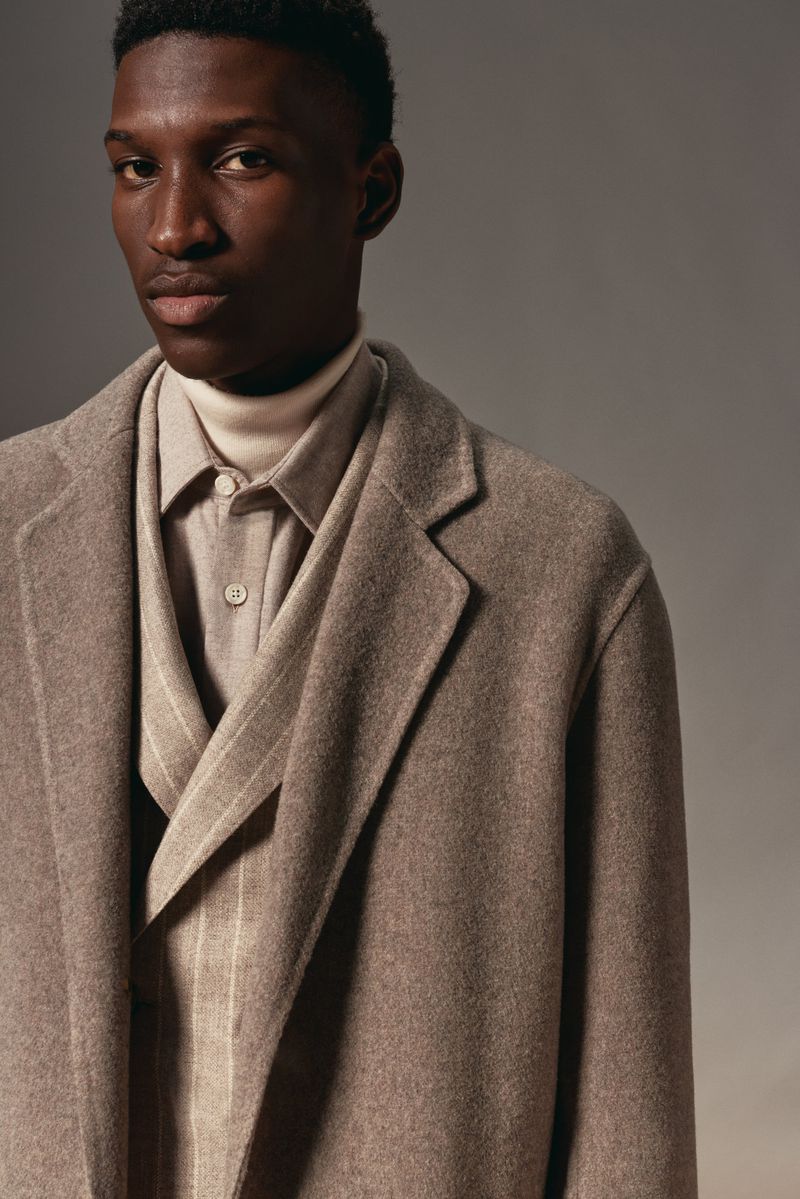 Portrait of a Black man in a stylish brown overcoat, ivory turtleneck, dress shirt, and vest against a neutral background.