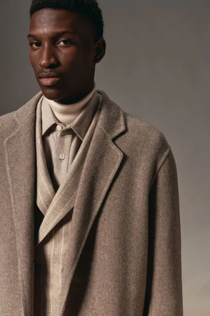 Portrait of a Black man in a stylish brown overcoat, ivory turtleneck, dress shirt, and vest against a neutral background.