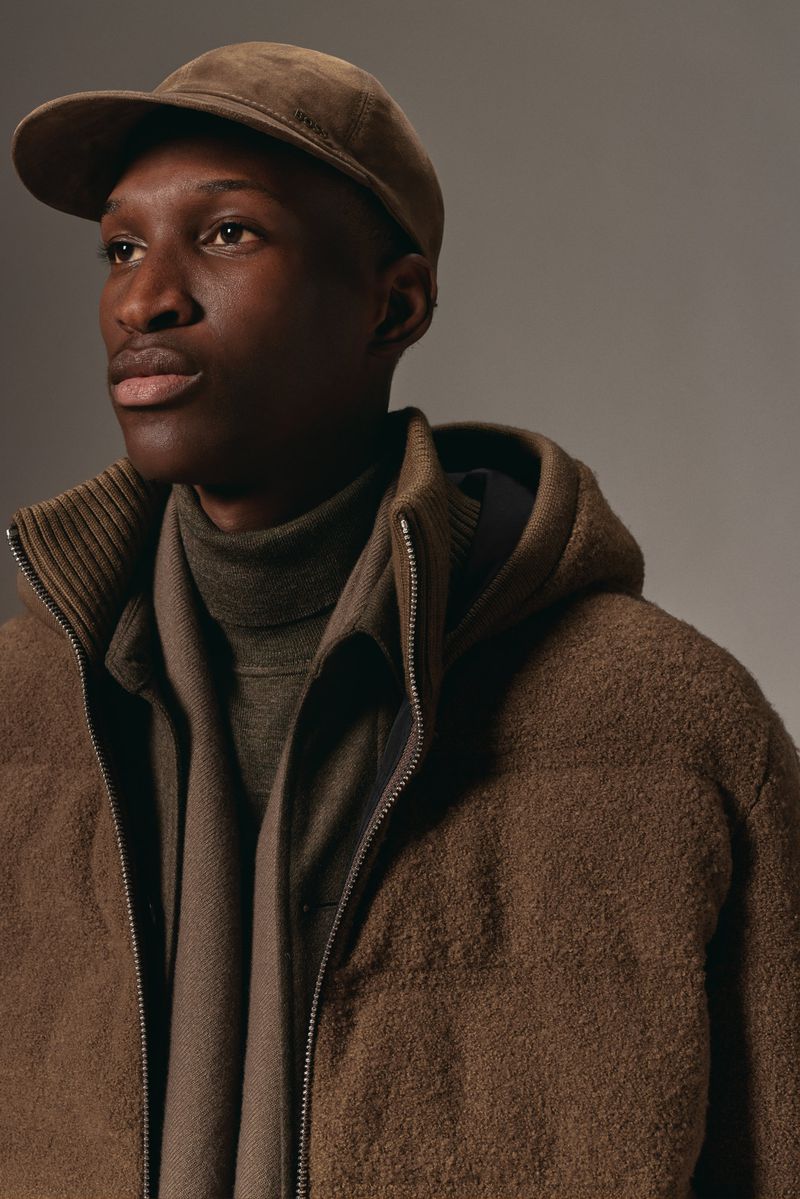 Portrait of a dark-skinned man wearing a cap, wool zip-up jacket, and a turtleneck sweater in a neutral studio setting with soft lighting.