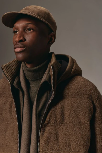 Portrait of a dark-skinned man wearing a cap, wool zip-up jacket, and a turtleneck sweater in a neutral studio setting with soft lighting.