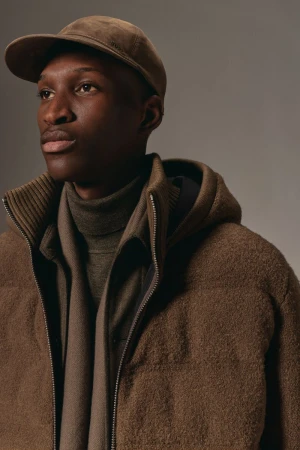 Portrait of a dark-skinned man wearing a cap, wool zip-up jacket, and a turtleneck sweater in a neutral studio setting with soft lighting.
