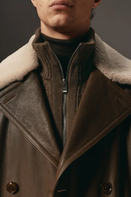 Close-up of a brown leather overcoat with a shearling collar, worn over a zip-up knit sweater, against a neutral grey backdrop.