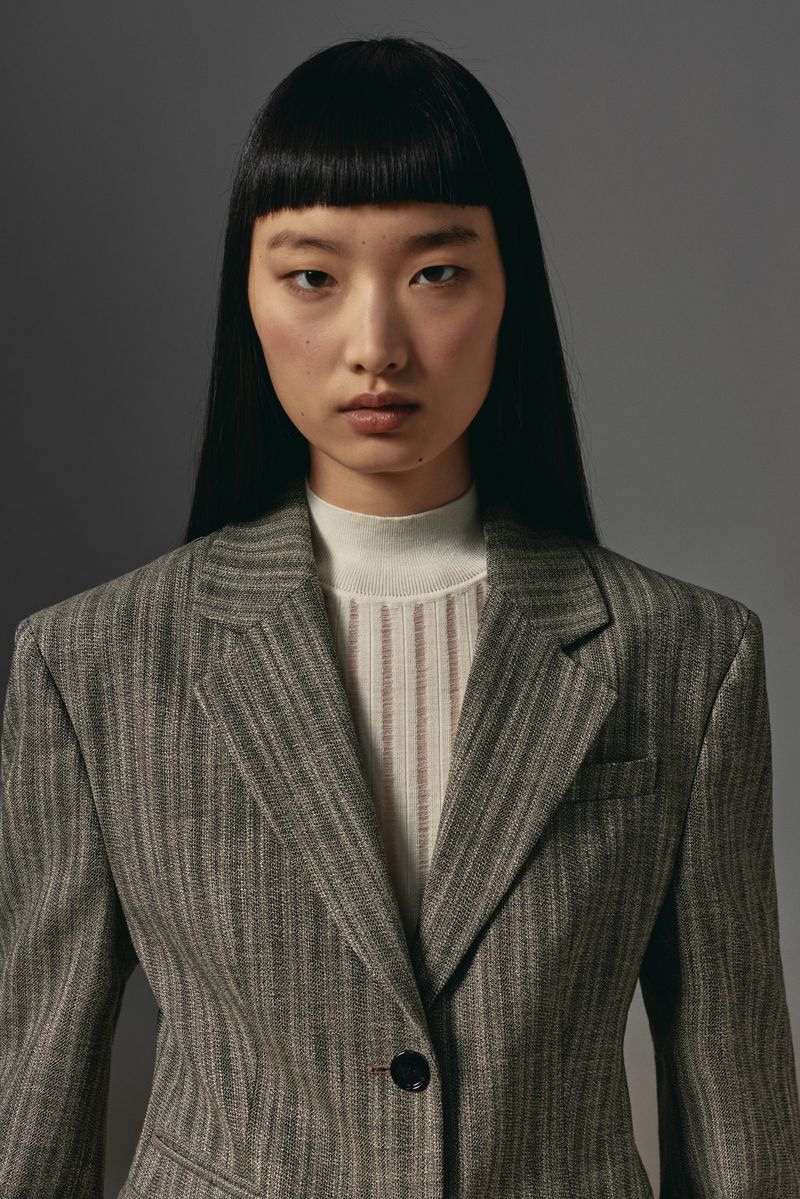 Close-up of a young Asian woman in a striped blazer, standing indoors against a grey background, exuding a sophisticated and modern style.
