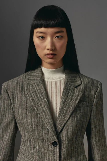 Close-up of a young Asian woman in a striped blazer, standing indoors against a grey background, exuding a sophisticated and modern style.
