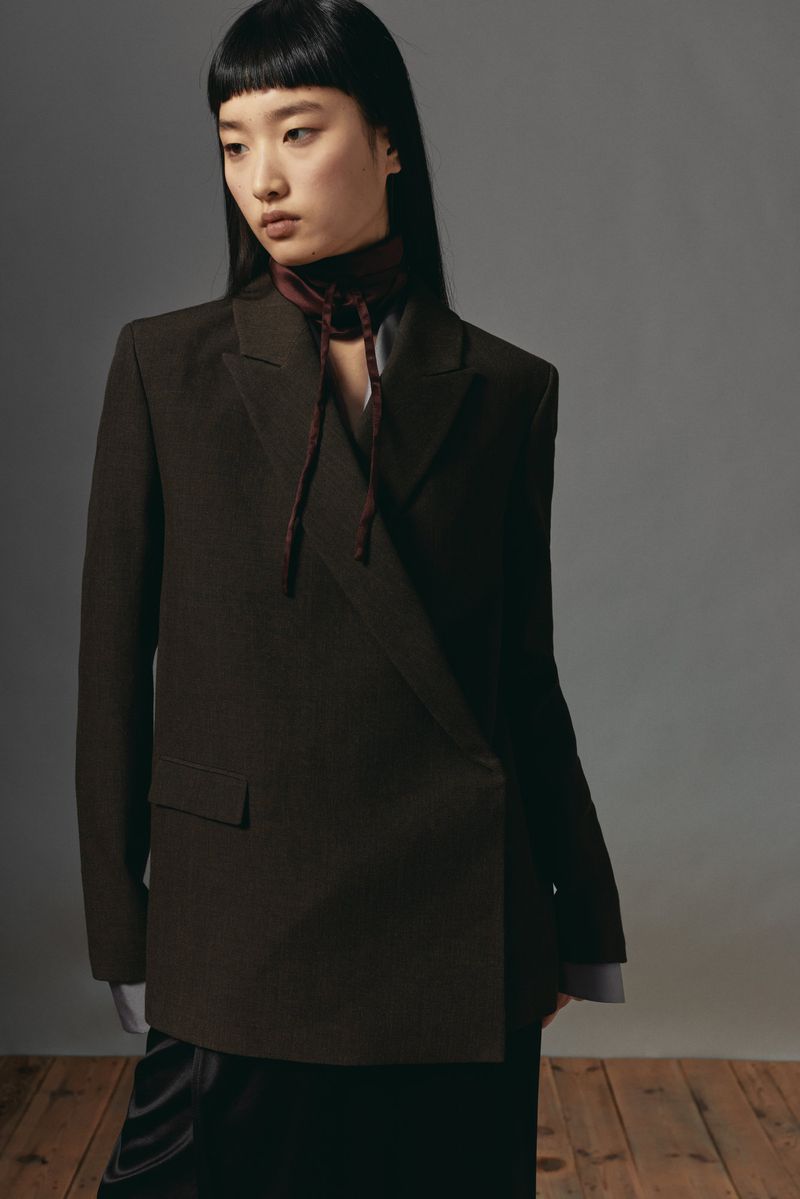 Model with long dark hair wearing a dark brown blazer and tie neck scarf against a grey background.