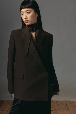 Model with long dark hair wearing a dark brown blazer and tie neck scarf against a grey background.