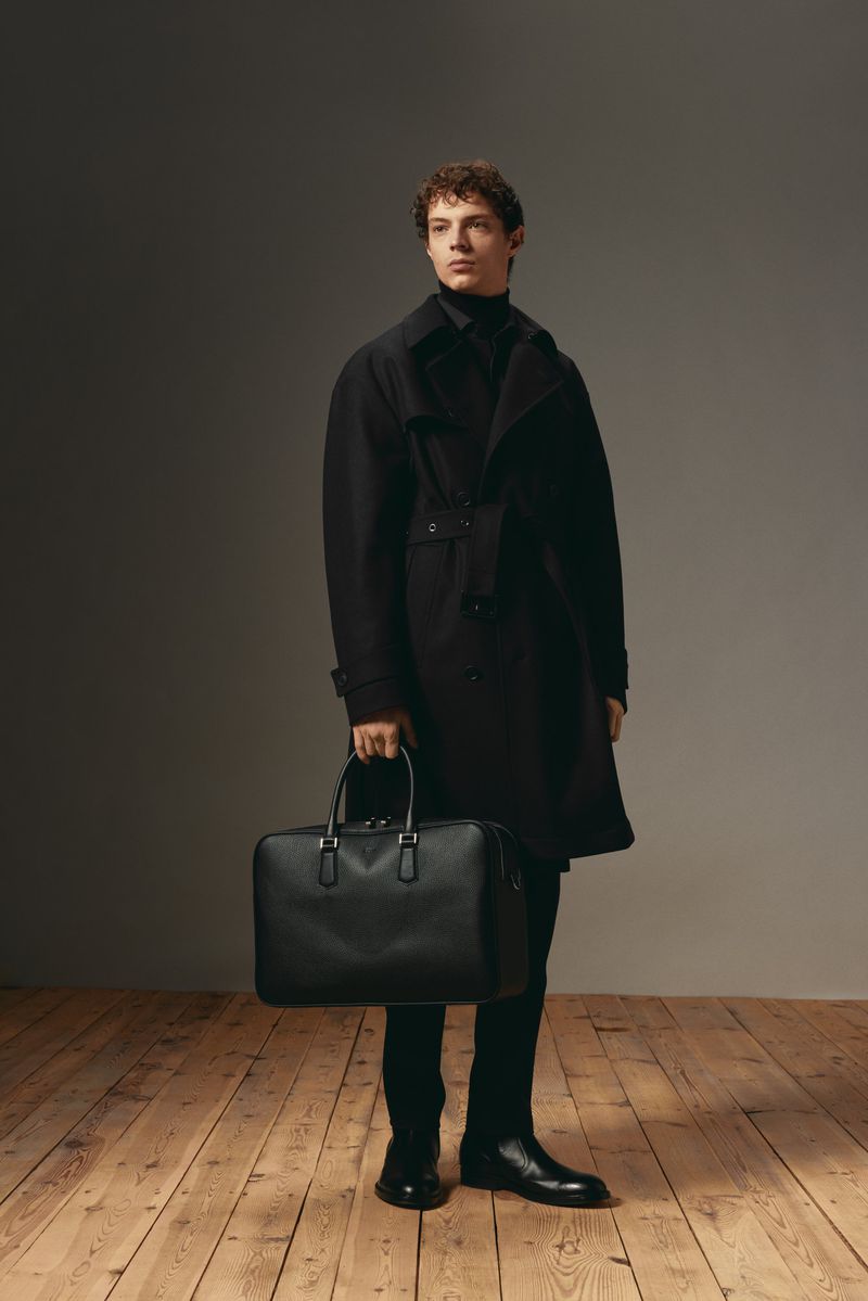 A stylish man stands in a studio, wearing a black turtleneck, coat, pants, and leather boots, holding a black leather briefcase.