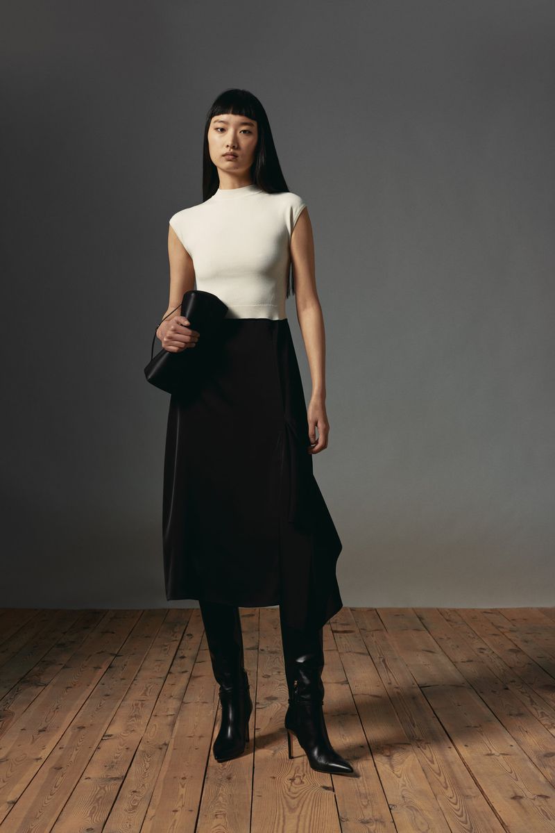 An Asian woman poses indoors wearing a cream-colored top, a dark skirt, high-heeled boots, and holding a dark-colored handbag.