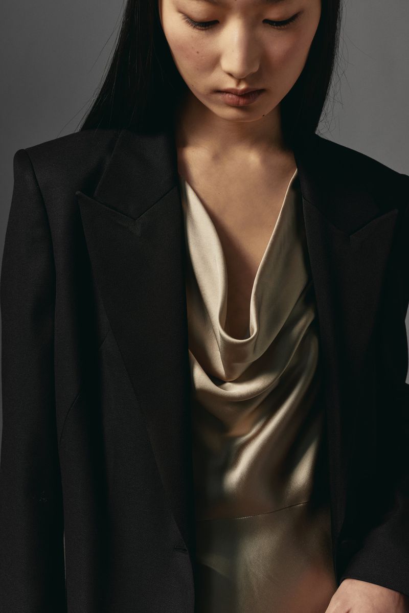 Asian woman in black blazer and cream cowl-neck dress posing against gray backdrop in studio, highlighting fashion and portrait photography.