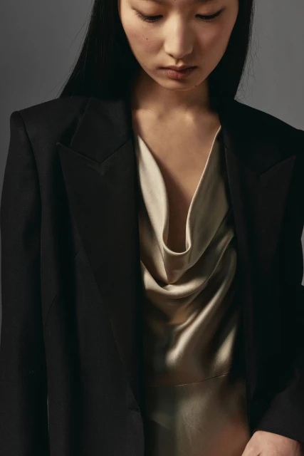 Asian woman in black blazer and cream cowl-neck dress posing against gray backdrop in studio, highlighting fashion and portrait photography.