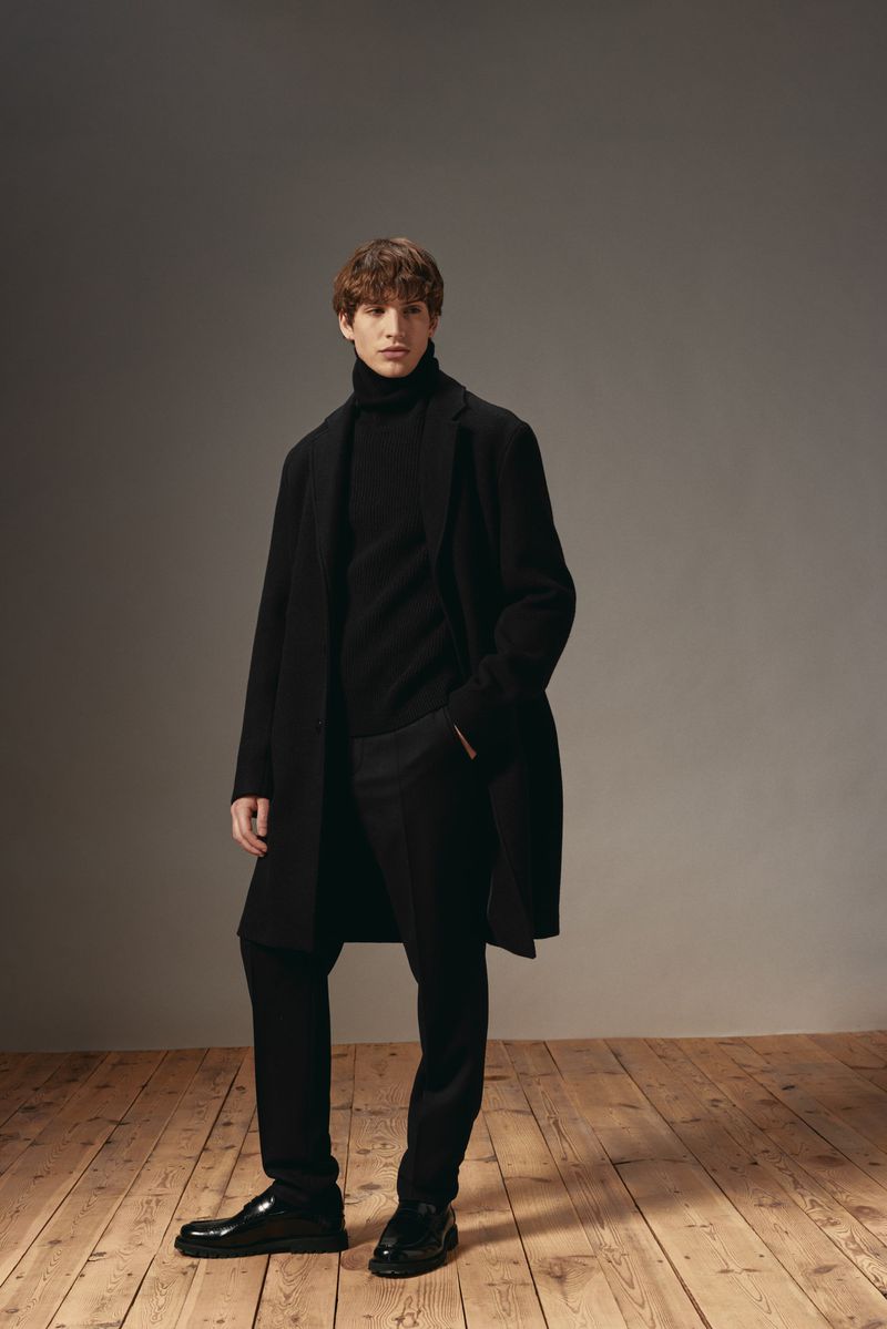 A young adult male model poses in a black turtleneck, black pants, black overcoat, and black shoes in a studio setting.