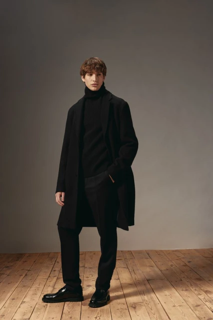 A young adult male model poses in a black turtleneck, black pants, black overcoat, and black shoes in a studio setting.