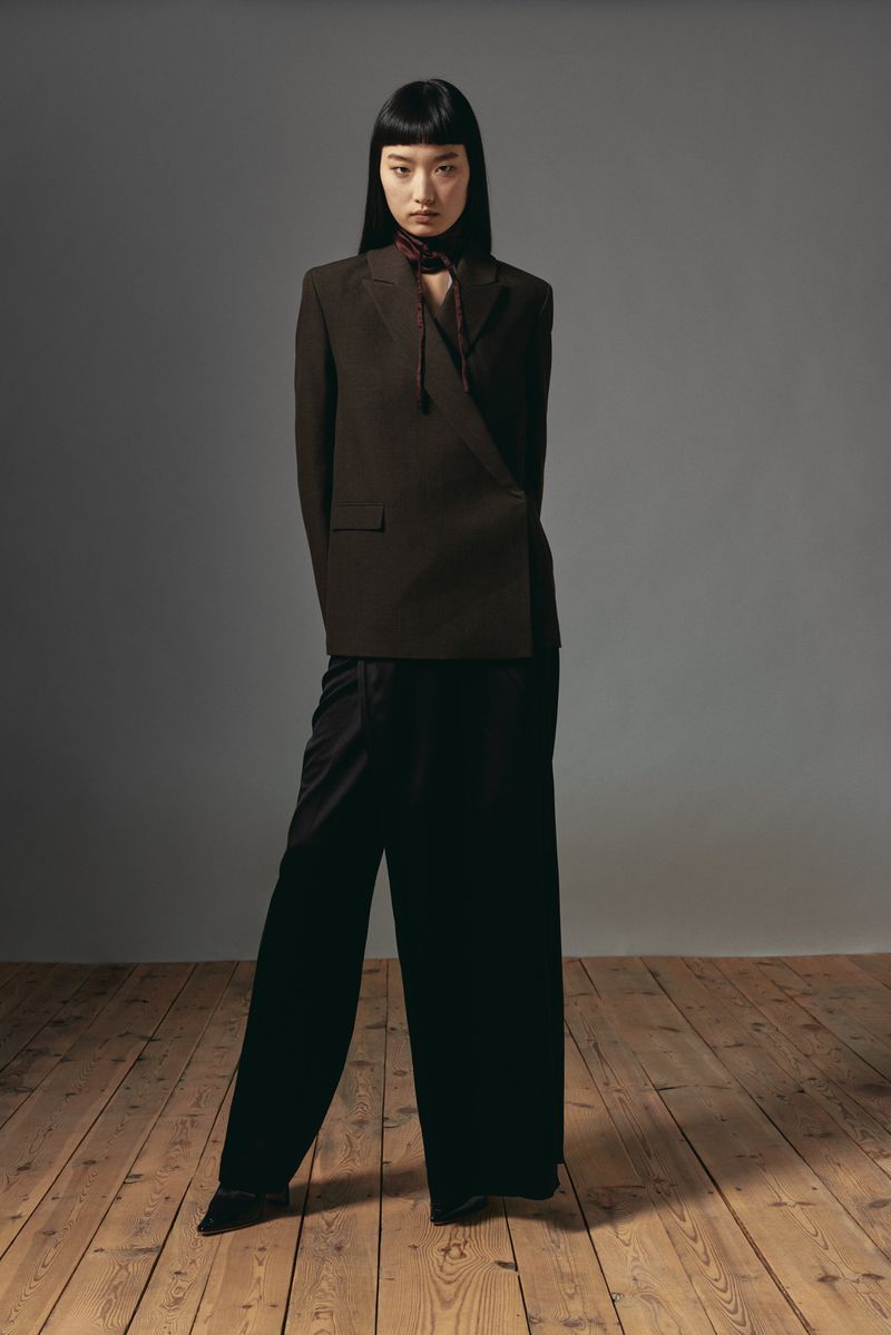 An adult woman poses in a studio wearing a brown blazer and black pants. She has dark hair with bangs and stands on a wood floor.