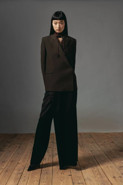 An adult woman poses in a studio wearing a brown blazer and black pants. She has dark hair with bangs and stands on a wood floor.