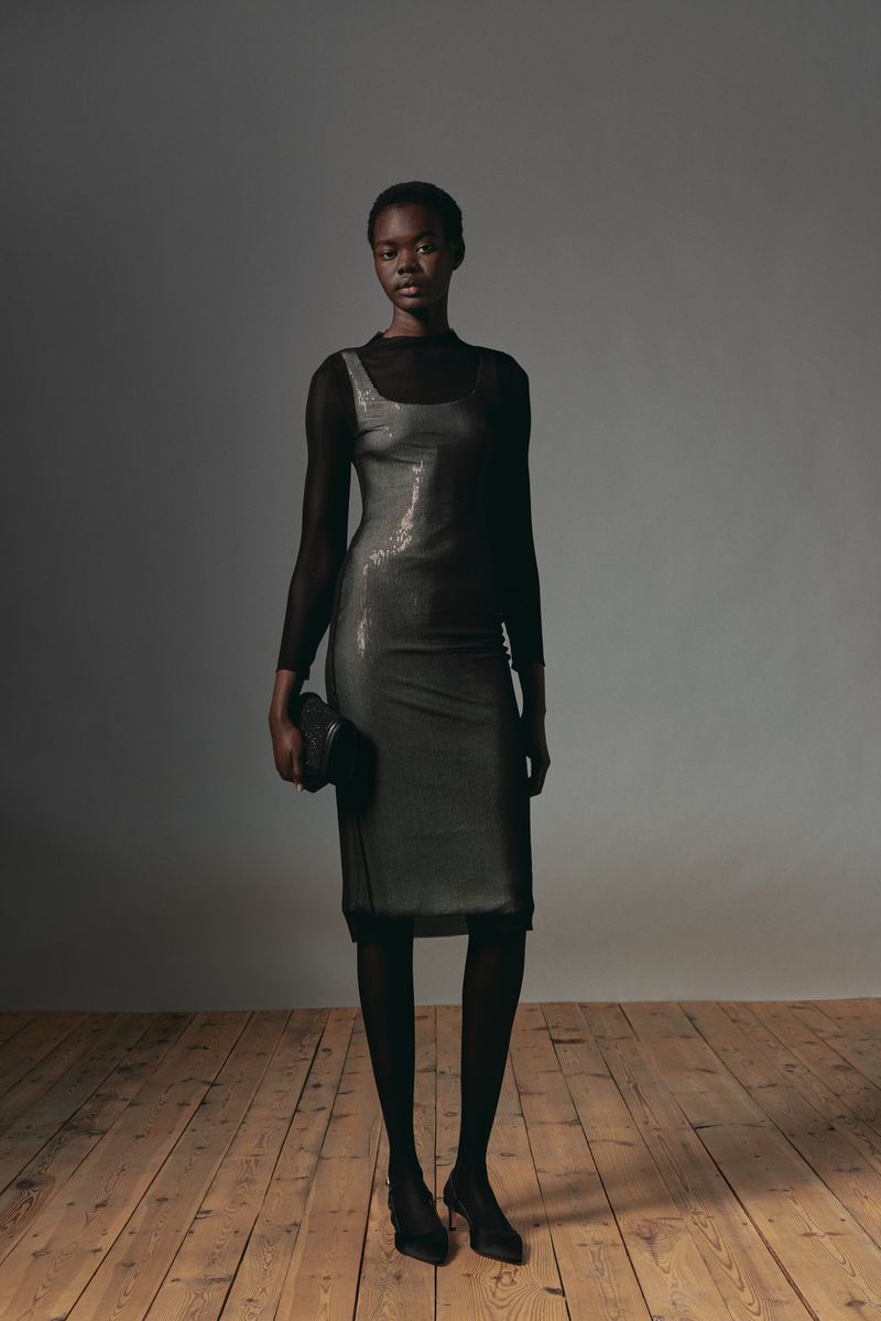 Full-length portrait of a Black female model in a black sequin dress, sheer black top, and black tights, holding a black clutch.
