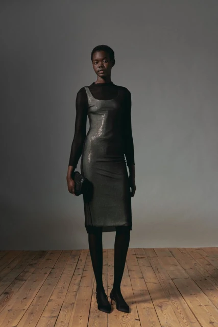 Full-length portrait of a Black female model in a black sequin dress, sheer black top, and black tights, holding a black clutch.