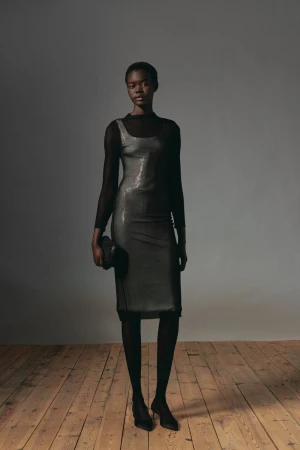 Full-length portrait of a Black female model in a black sequin dress, sheer black top, and black tights, holding a black clutch.