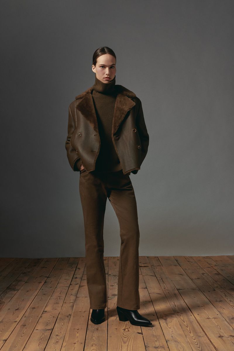 Model standing on a wooden floor wearing a brown shearling jacket and matching trousers against a grey wall