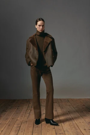 Model standing on a wooden floor wearing a brown shearling jacket and matching trousers against a grey wall