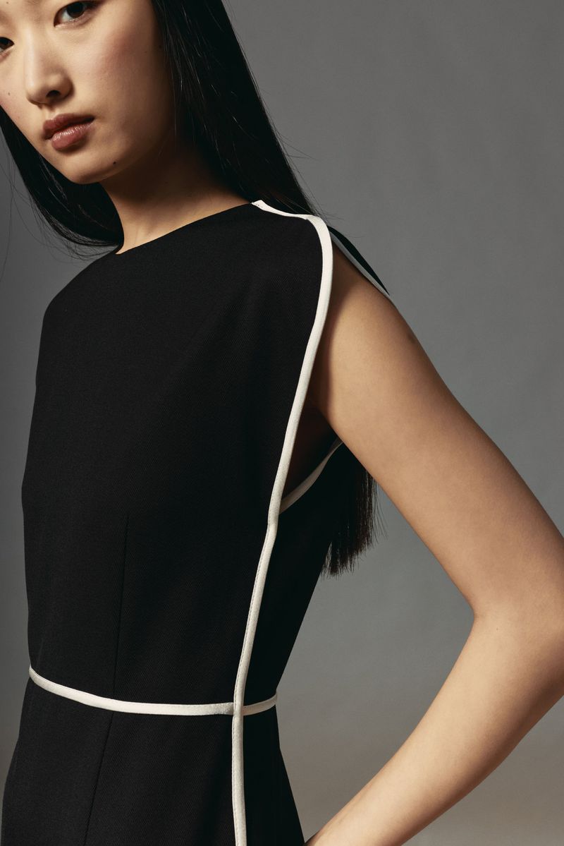 Studio shot of a slender woman with dark hair wearing a sleeveless black dress with white trim. Fashion, style, and simple elegance.