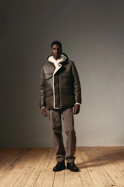 Full-length portrait of a young African-American male model posing in a brown shearling jacket and brown trousers, standing on wood flooring.