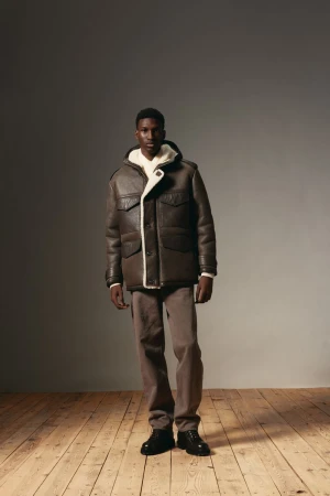 Full-length portrait of a young African-American male model posing in a brown shearling jacket and brown trousers, standing on wood flooring.