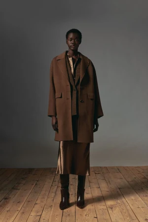 Model wearing a brown coat and satin skirt standing on wooden floorboards against a neutral grey studio wall.