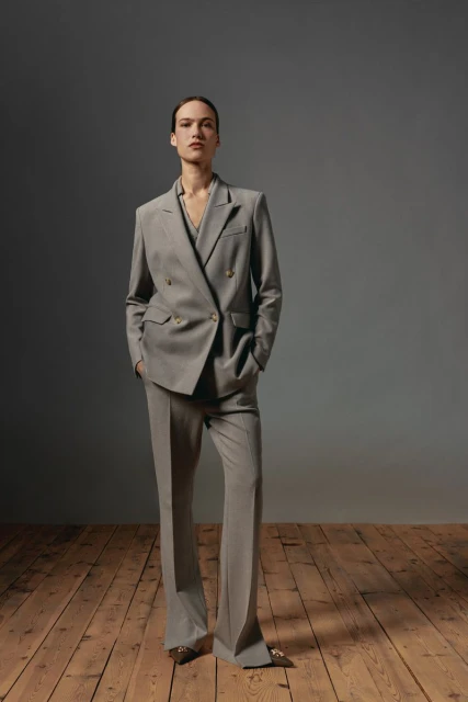 Adult woman in a grey double-breasted blazer and wide-leg pants stands in a studio with wood floors and a grey backdrop.