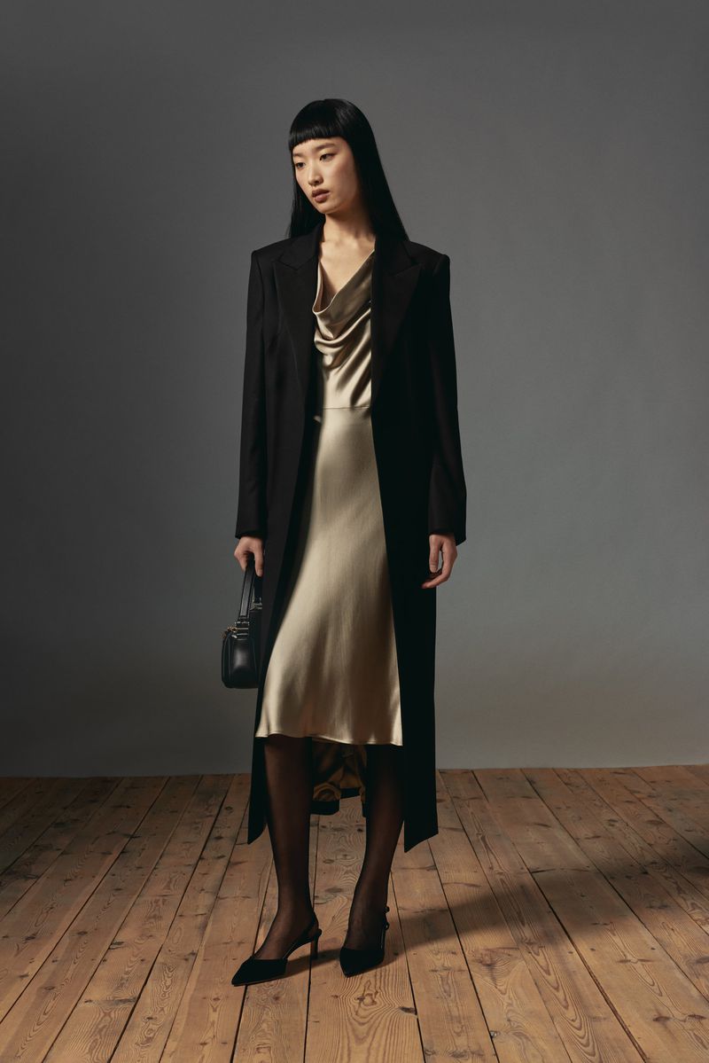 Model with long dark hair wearing a long black coat over a satin slip dress and holding a handbag on a wooden floor.