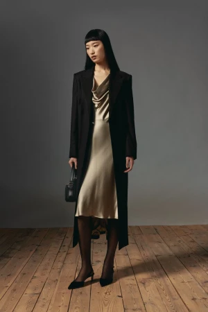 Model with long dark hair wearing a long black coat over a satin slip dress and holding a handbag on a wooden floor.