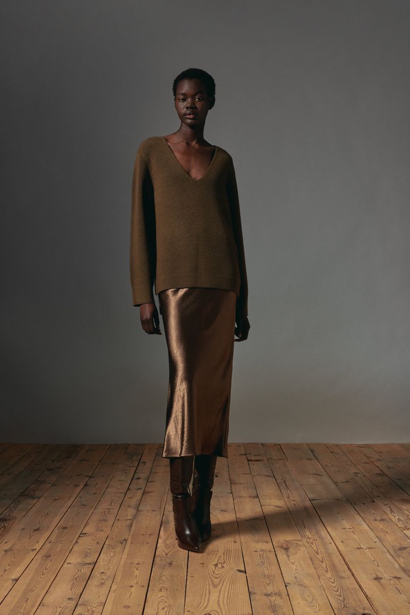 Elegant Black woman poses in a stylish green sweater, satin skirt, and knee-high boots on a wood floor against a grey backdrop.