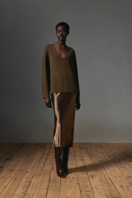 Elegant Black woman poses in a stylish green sweater, satin skirt, and knee-high boots on a wood floor against a grey backdrop.