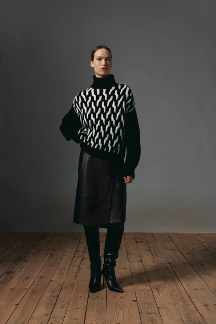 Full-length shot of a woman in a black and white patterned sweater, black leather skirt, leggings, and heeled boots on a wood floor.