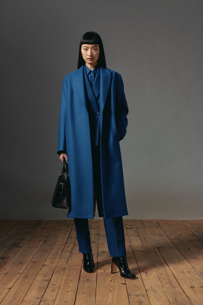Model wearing a blue coat and holding a black handbag standing on a wooden floor against a grey wall.