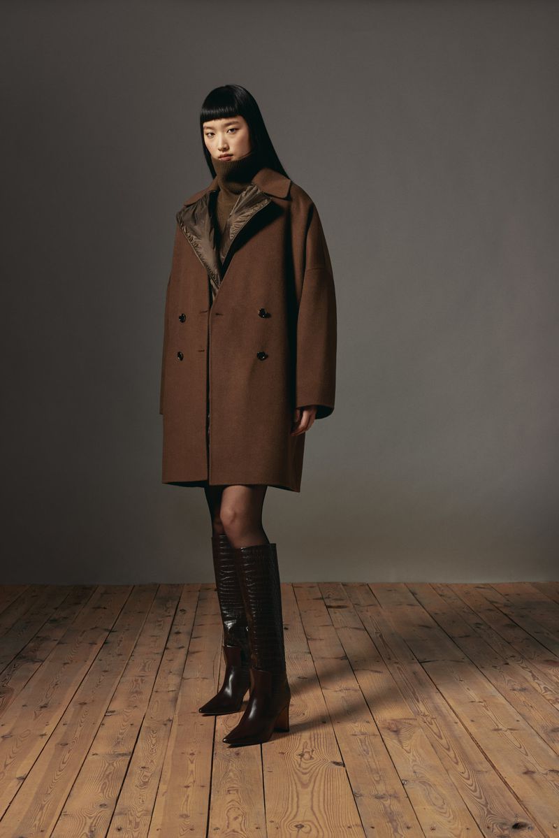 A stylish Asian woman poses in a brown double-breasted wool coat, brown turtleneck, sheer black tights, and brown leather knee-high boots.